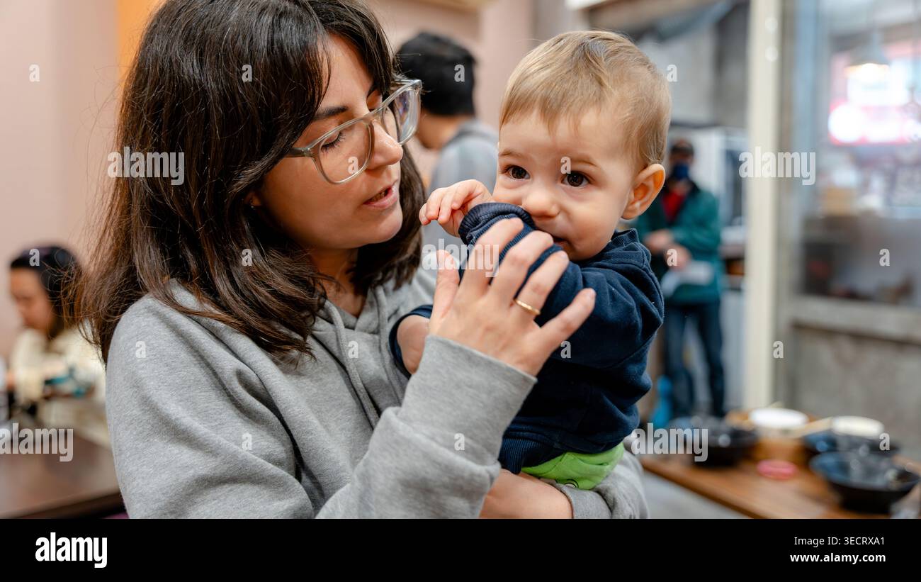 Una donna tiene in braccio un bambino mentre cena in un vivace ristorante taiwanese. L'atmosfera accogliente e l'arredamento tradizionale creano un ambiente accogliente e adatto alle famiglie Foto Stock