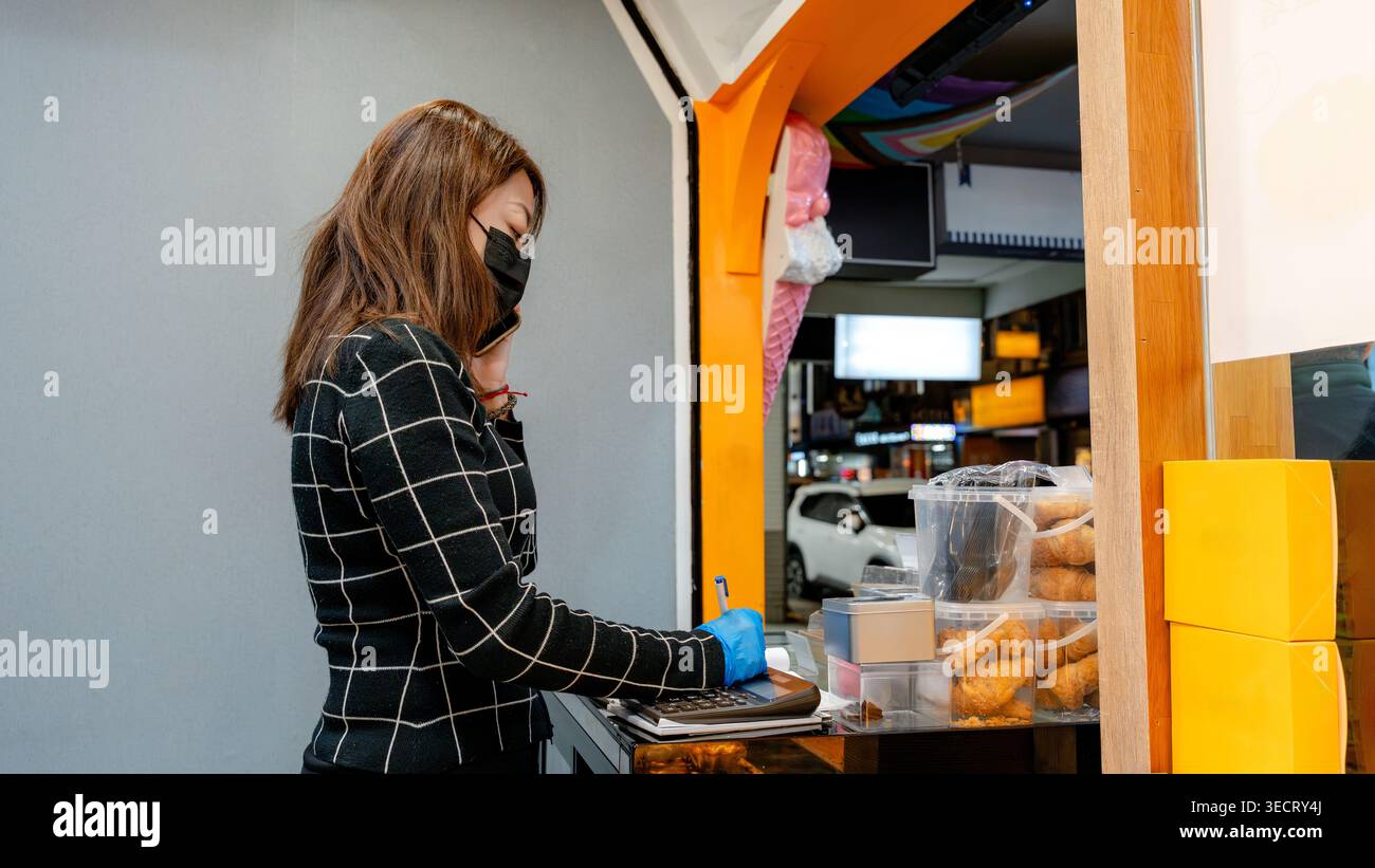 Una donna che indossa una maschera lavora in una panetteria di Taipei, organizzando gli oggetti e utilizzando una calcolatrice. L'ambiente luminoso offre uno sguardo sulla vivace atmosfera locale Foto Stock