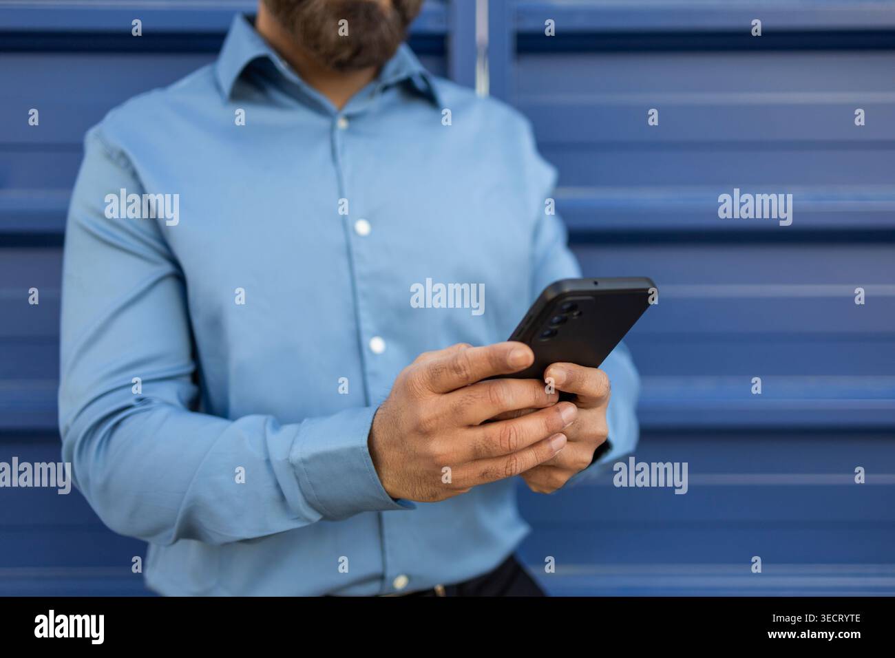 Vista ritagliata di un uomo d'affari con una camicia blu che regge uno smartphone, legge messaggi e gestisce il lavoro mobile, in piedi contro un otturatore blu in metallo b Foto Stock