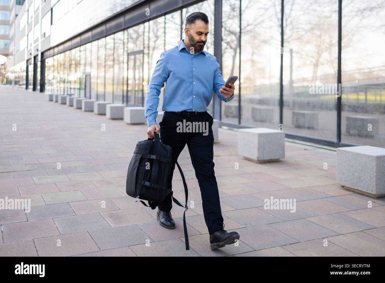 Un uomo d'affari arabo in camicia blu cammina lungo un moderno edificio per uffici, porta con sé uno zaino e controlla il suo smartphone, catturando gli spostamenti urbani, mobil Foto Stock