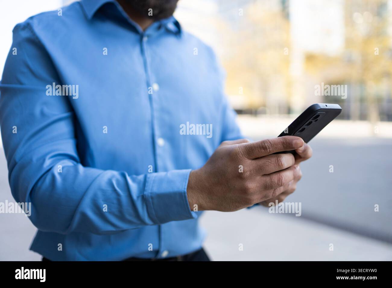 Primo piano di un uomo d'affari con una camicia blu che regge uno smartphone all'aperto, controlla i messaggi e gestisce le attività di lavoro mobili, la comunicazione digitale e la Foto Stock