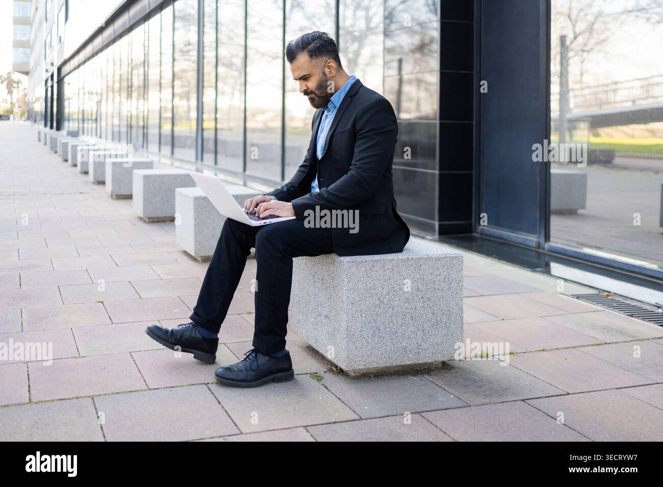 Un uomo d'affari arabo in tuta nera siede su una panchina di pietra all'esterno di un moderno edificio di uffici in vetro e utilizza un computer portatile per lavorare in remoto, pianificare e co-on-line Foto Stock