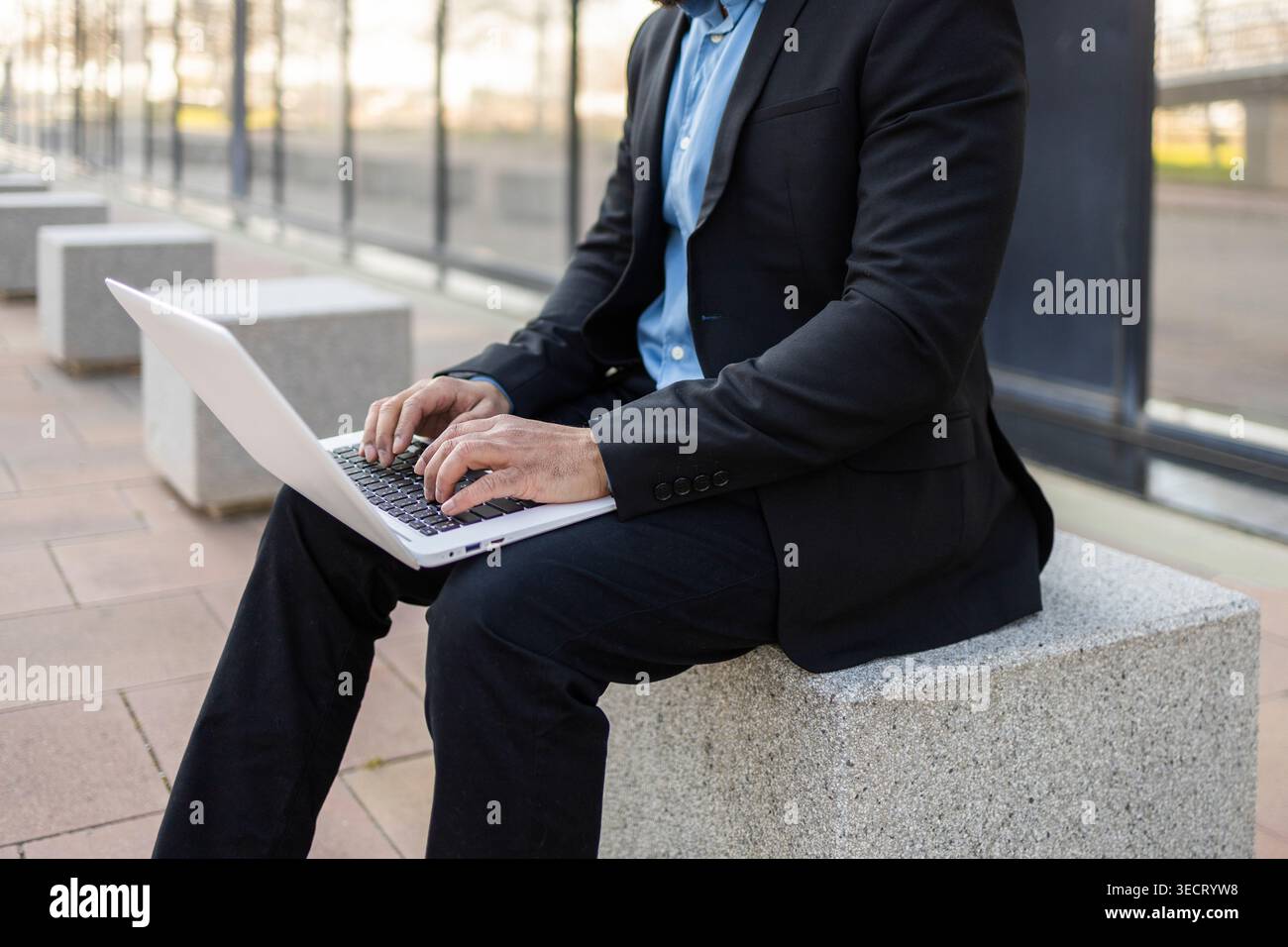 Uomo d'affari con una tuta nera che utilizza un computer portatile mentre è seduto su una panca di cemento all'aperto accanto a un moderno edificio per uffici, concentrato su lavoro remoto, e-mail, a. Foto Stock