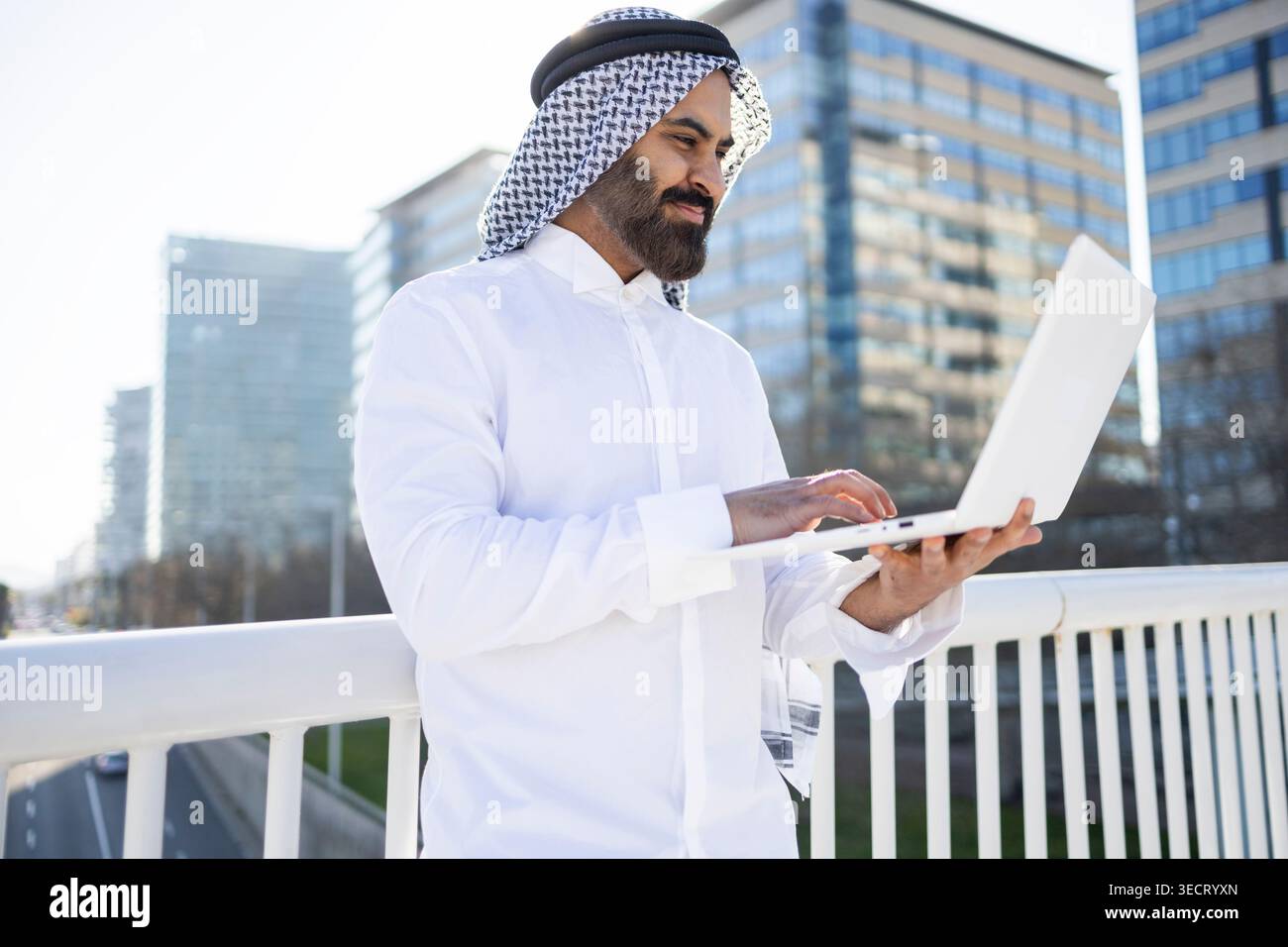 Uomo d'affari arabo in tradizionale abbigliamento sheikh che utilizza un laptop all'aperto vicino ai moderni uffici di vetro, lavora in remoto con fiducia su un luminoso urb Foto Stock