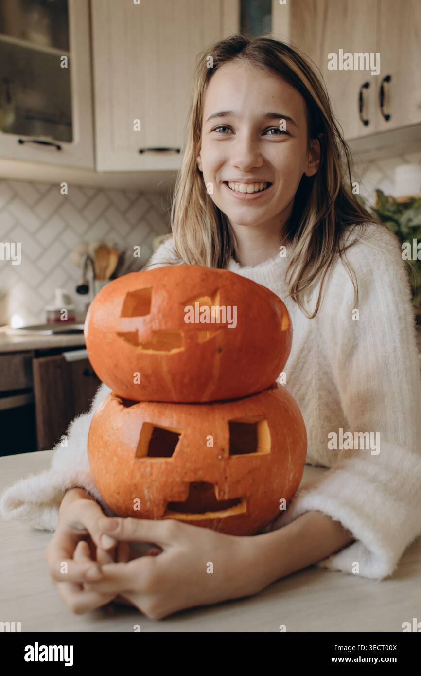 Ragazza sorridente in casa che tiene in mano le zucche scolpite di Halloween su un tavolo da cucina, atmosfera accogliente con decorazioni autunnali, facce di Jack-o'-lantern e Foto Stock