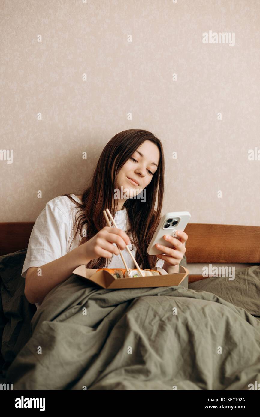 Una donna ama trascorrere una giornata rilassante a casa, sedersi comodamente a letto mentre mangia sushi e naviga sul suo smartphone, per una giornata perfetta e accogliente Foto Stock