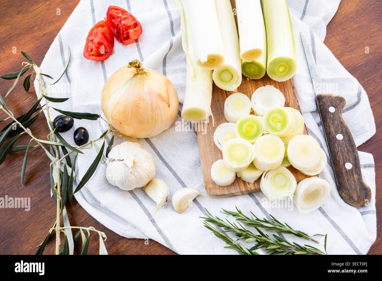 Vista dall'alto di porri freschi tagliati su un asse di legno con cipolla, spicchi d'aglio, pomodori, rametti d'oliva e rosmarino su un asciugamano da cucina a righe, leggere Foto Stock