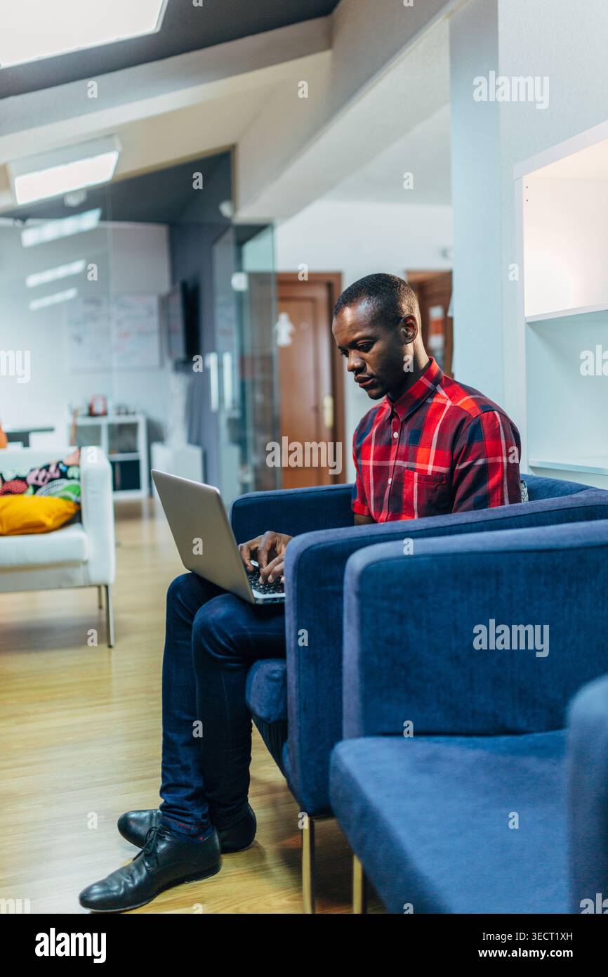 Un giovane uomo Nero con una camicia a quadri rossa si trova su un divano blu, scrivendo su un computer portatile in una luminosa e moderna sala ufficio con pareti in vetro e un comodo sedile Foto Stock
