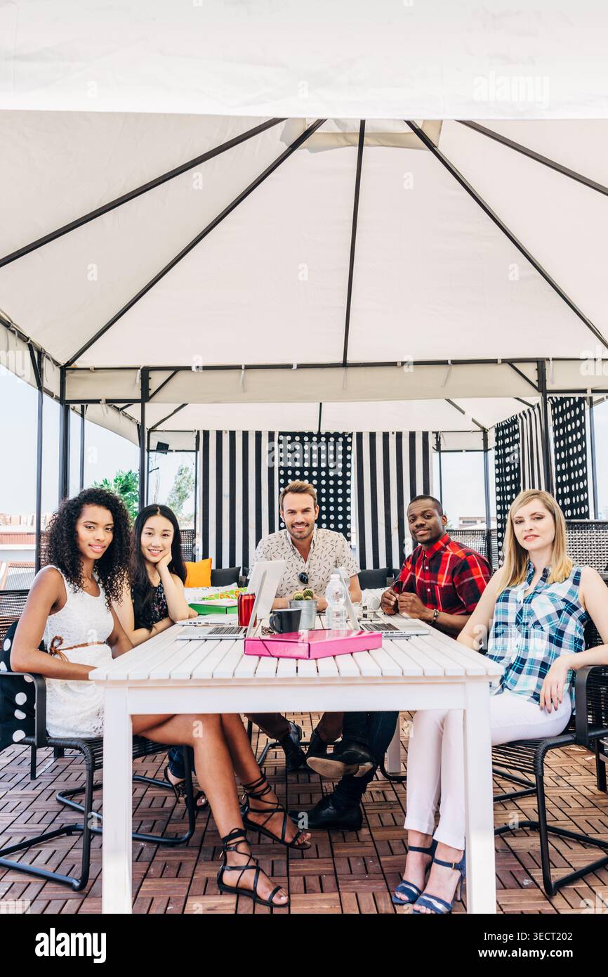 Cinque diversi giovani adulti si siedono insieme a un tavolo sul patio sul tetto sotto un gazebo, sorridendo e chiacchierando sorseggiando un drink, godendosi un'informale riunione estiva Foto Stock