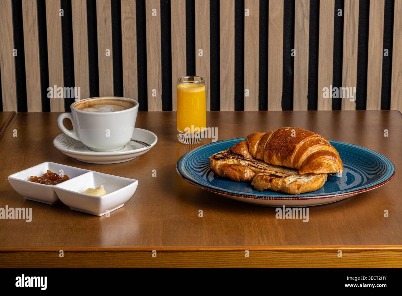 Colazione mattutina su un tavolo di legno con croissant, caffè caldo in una tazza bianca, succo d'arancia e piccole ciotole di marmellata e burro in un accogliente caffè Foto Stock
