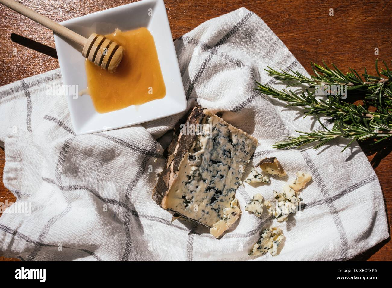 Primo piano di cremoso formaggio blu con briciole accanto a un piccolo piatto di miele e cucchiaio, stilato su un panno rustico con rametti di rosmarino in una tiepida ligine Foto Stock
