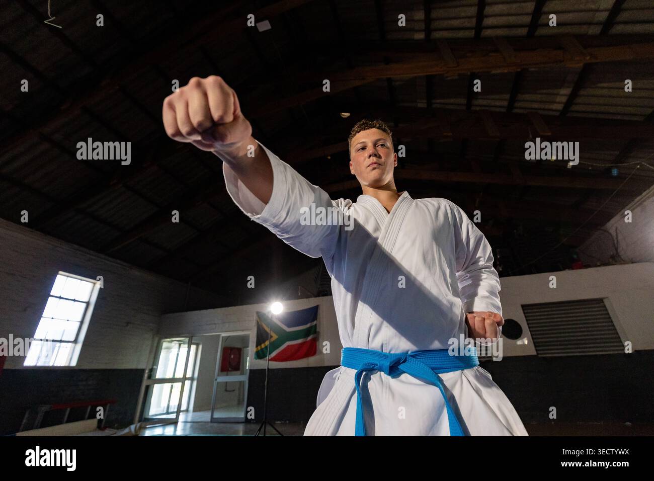 Uno studente di karate che indossa una cintura bianca e blu dà un forte colpo in un'ampia sala da allenamento. Illuminazione a basso angolo e ampia Foto Stock