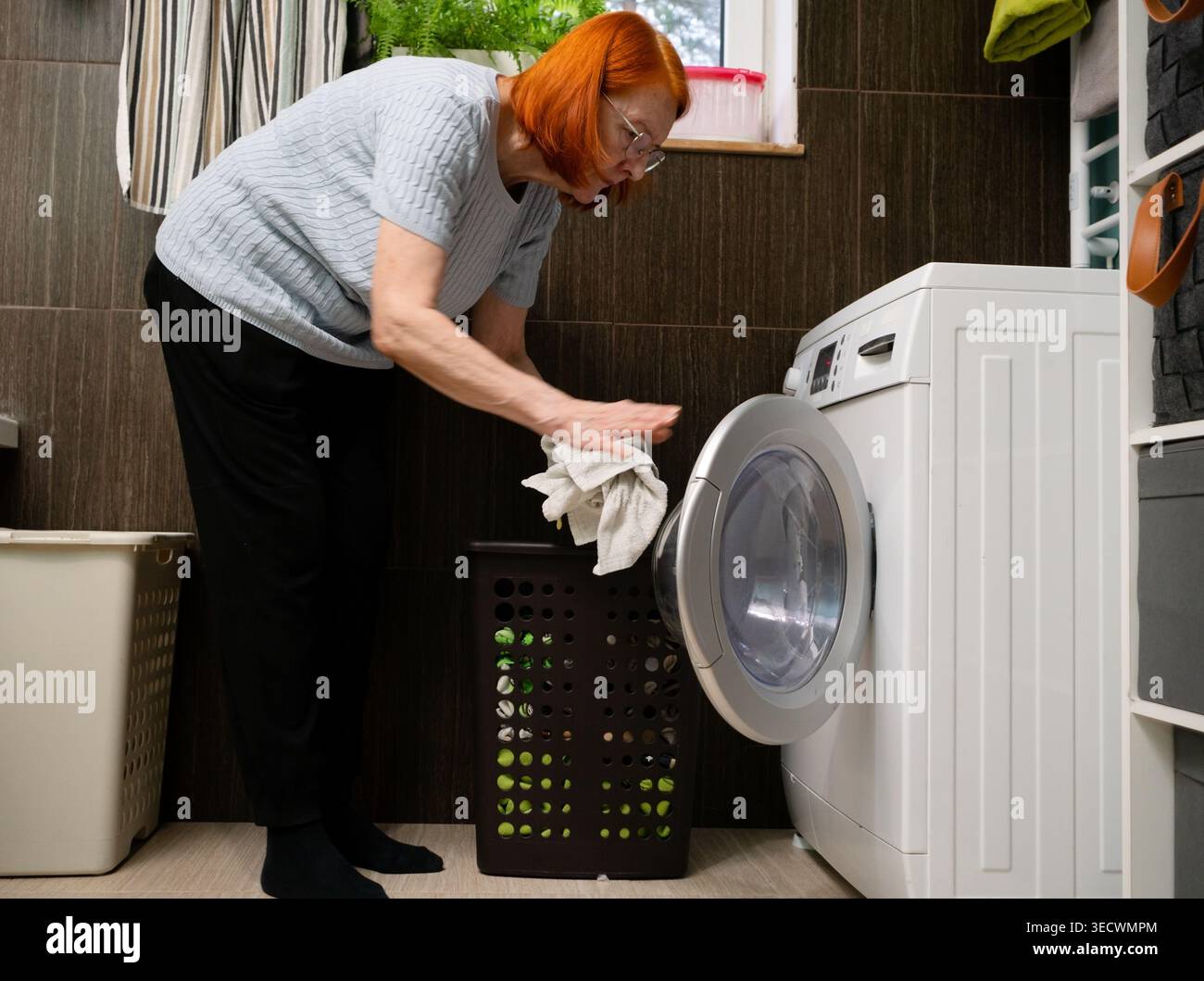 Una donna anziana aggiunge gli articoli finali dal cestino nella lavatrice, completando il processo di caricamento prima del lavaggio. Foto Stock