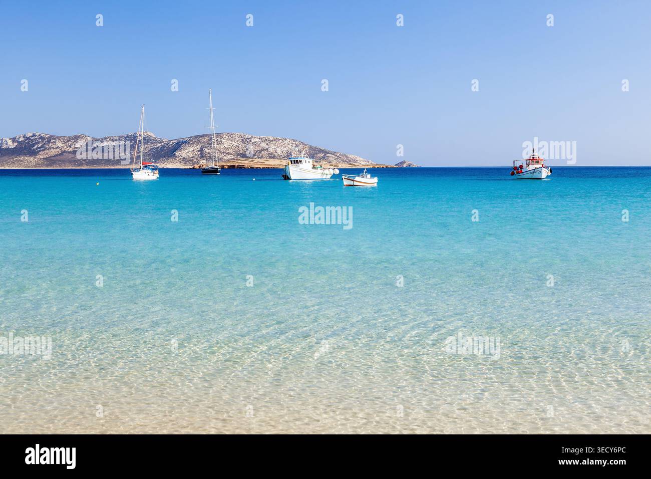 Mare cristallino e turchese con barche ancorate al largo dell'isola di Koufonisi, in Grecia, visto dalla costa sabbiosa poco profonda con le aspre isole cicladi sul retro Foto Stock