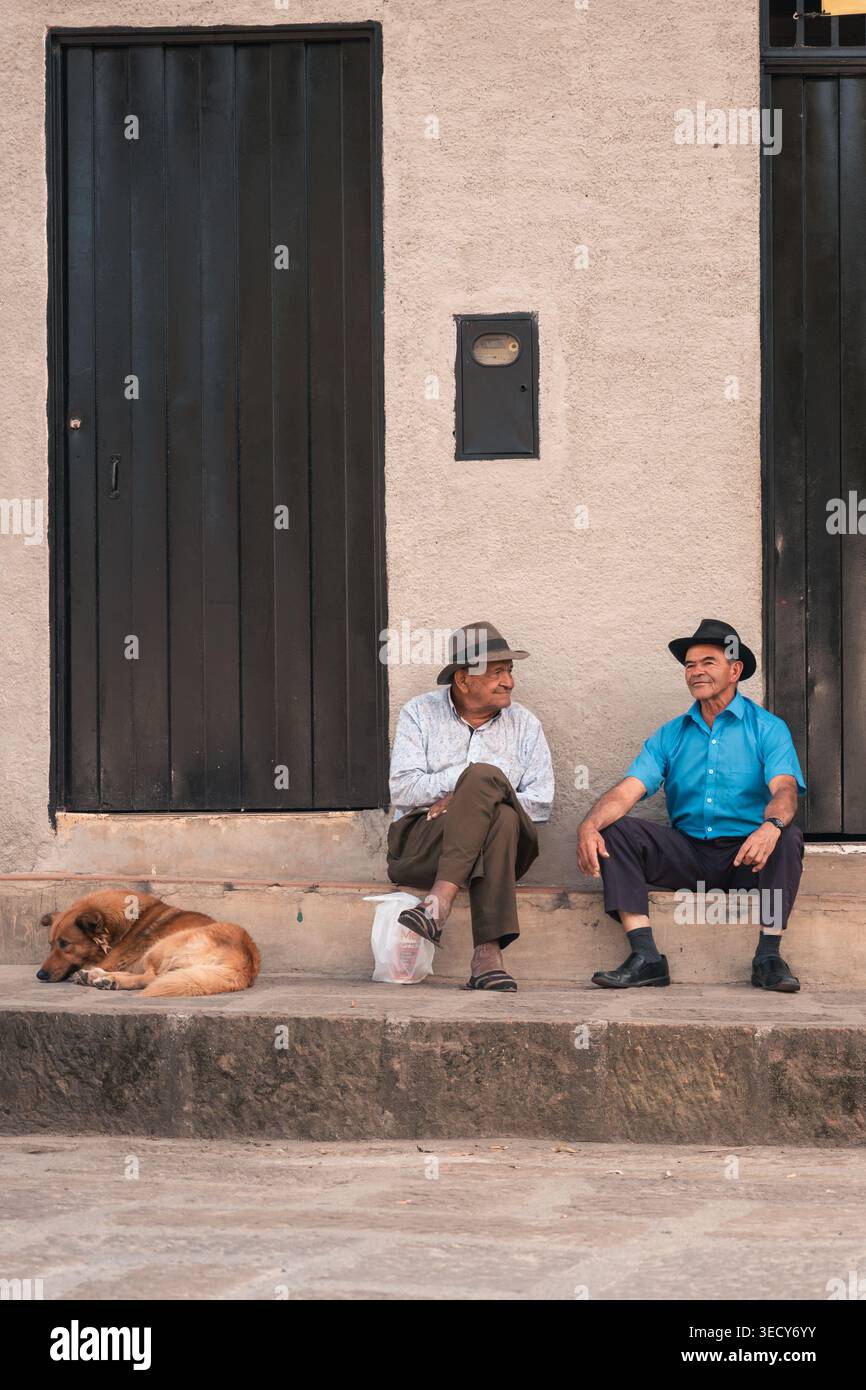 Mogotes, colombia - 21 marzo 2026 due uomini più anziani in cappello si siedono e chiacchierano in una strada soleggiata vicino a un edificio, un cane che riposa nelle vicinanze, scena del quartiere a Los Angeles Foto Stock