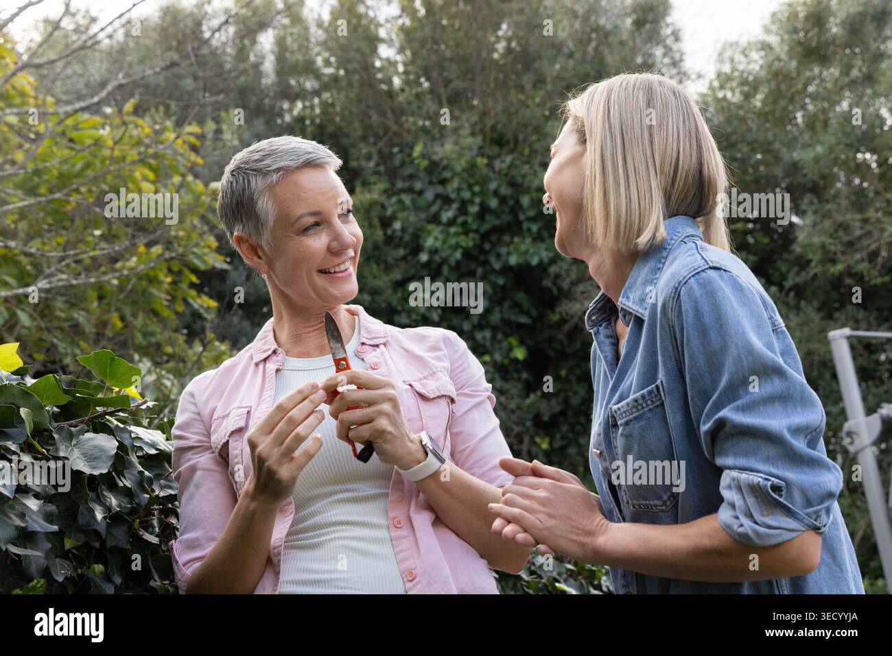 Donne anziane e di mezza età in piedi nel cortile con camicia in denim che regge cesoie per potare ridendo Foto Stock