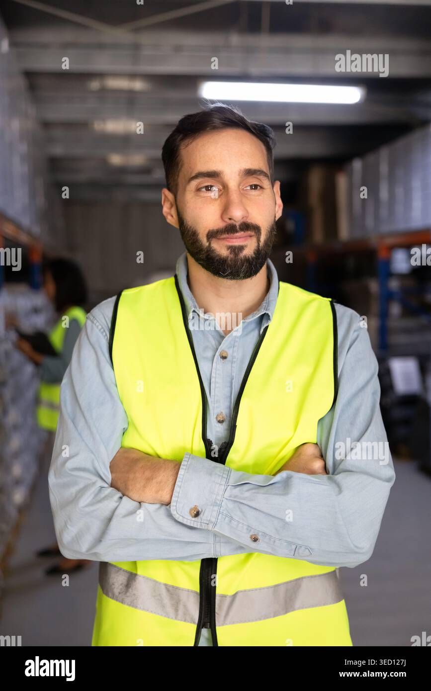Il lavoratore di magazzino indossa un giubbotto di sicurezza, sta con le braccia incrociate nel magazzino Foto Stock
