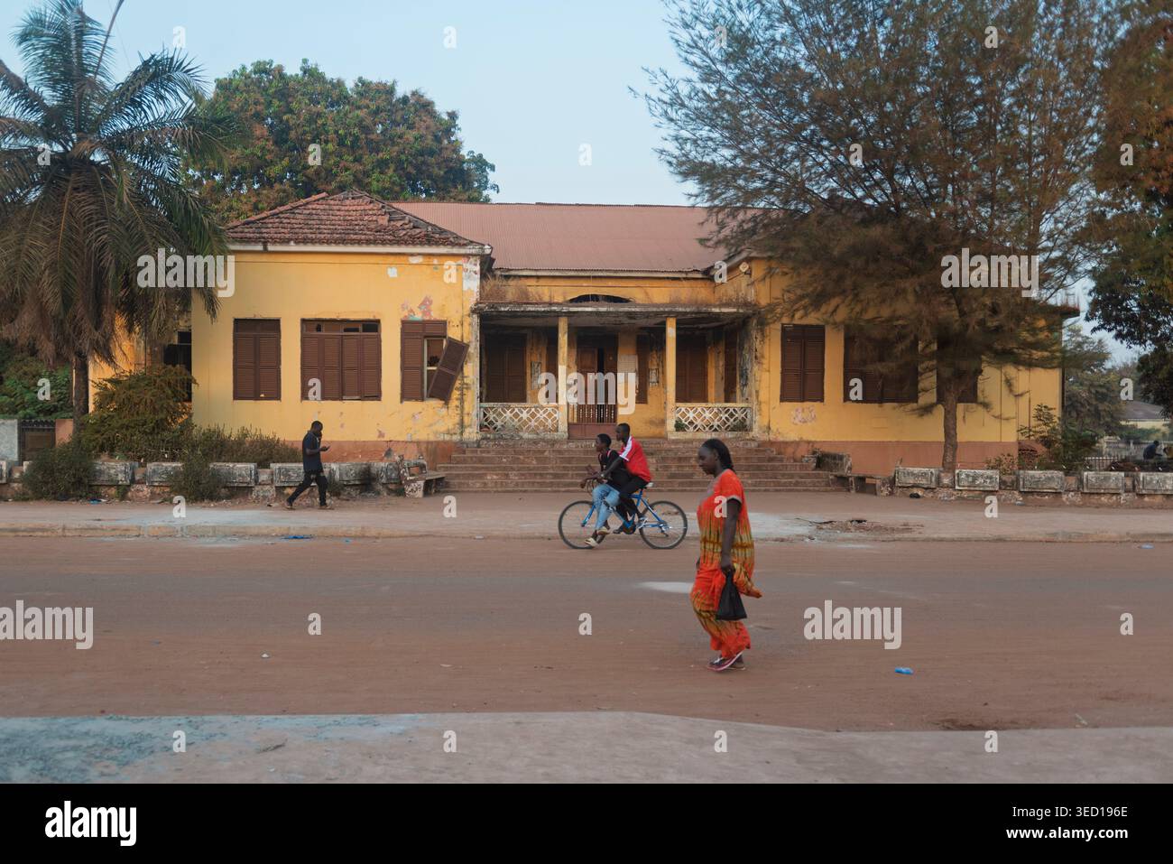 Donna in abito rosso e due adolescenti in bicicletta passano davanti a una villa coloniale color ocra fatiscente a Gabu, Guinea Bissau, Africa occidentale. Foto Stock