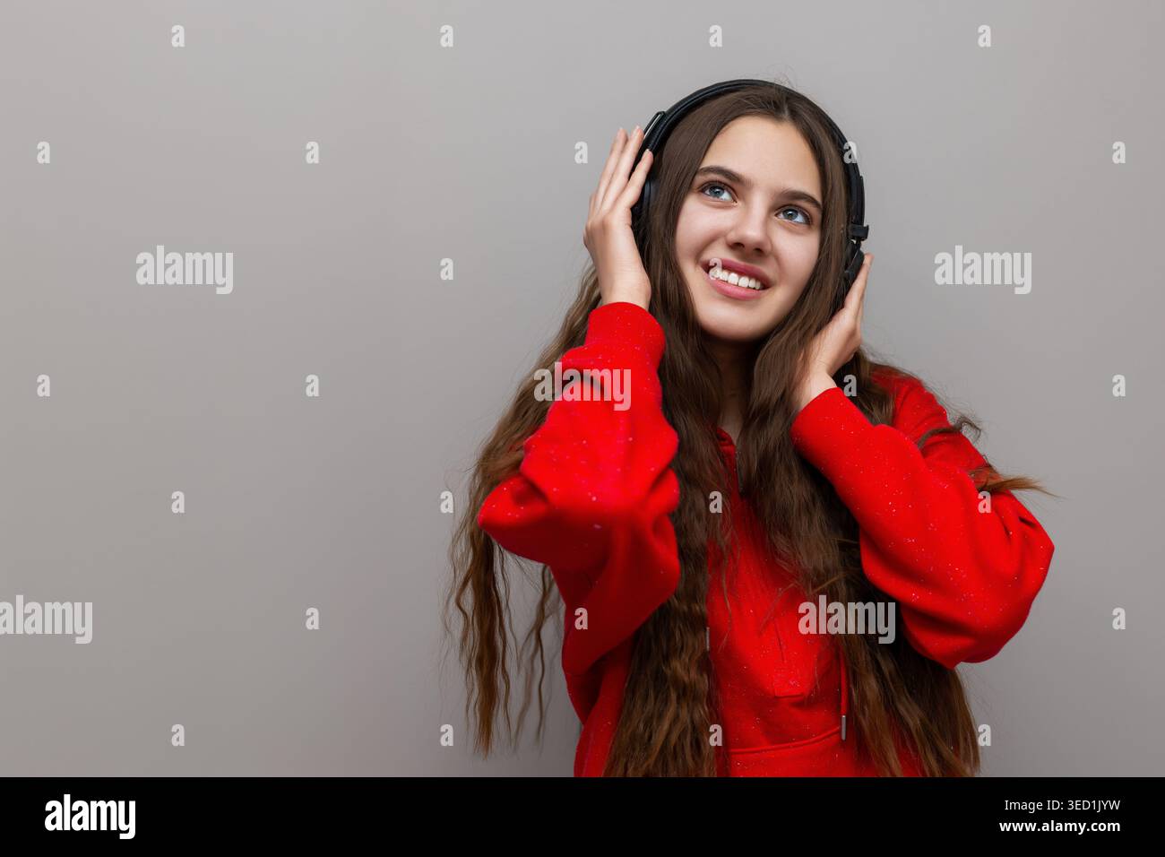 Felice giovane donna che ascolta la musica. Una giovane donna sorridente con cappuccio rosso ama la musica attraverso le cuffie, con un aspetto allegro e un semplice grigio Foto Stock