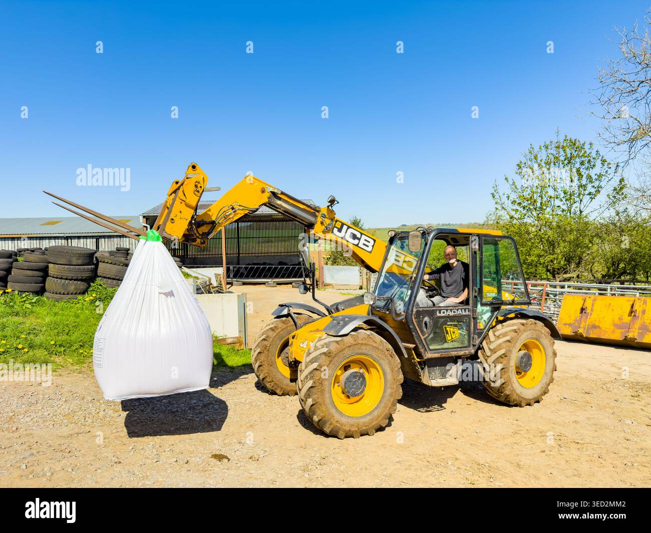 Movimentatore telescopico JCB giallo che solleva un grande sacchetto bianco in un'azienda agricola sotto un cielo azzurro Foto Stock