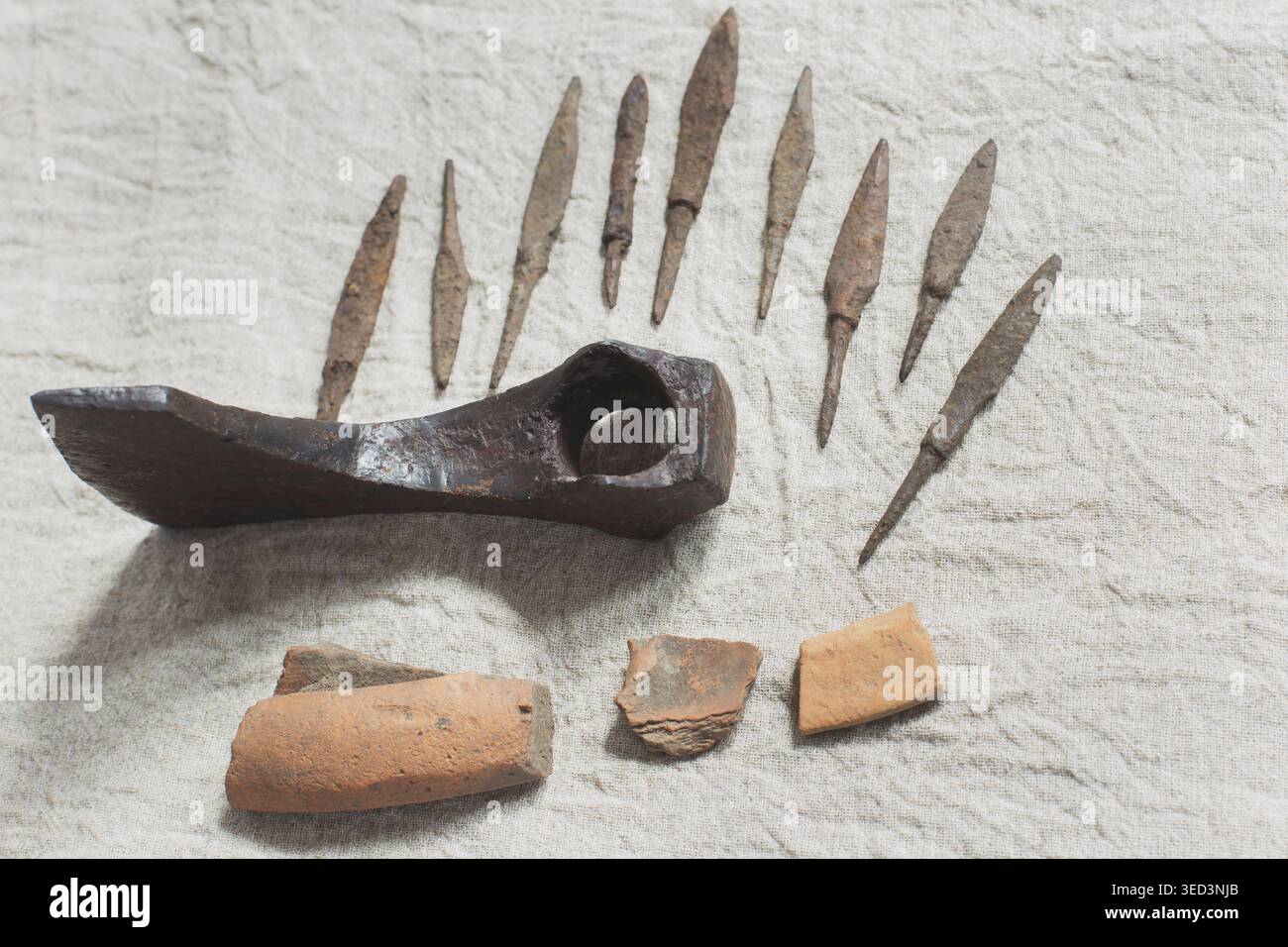 Antica testa di ascia di ferro arrugginita, punte di freccia e frammenti di ceramica su un fondo di lino naturale. Foto Stock