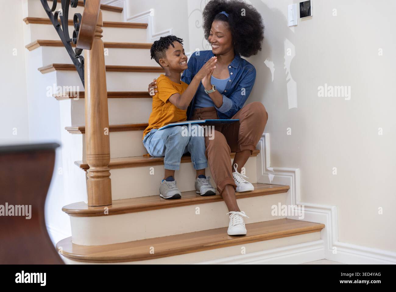 Madre afroamericana e figlio ispanico seduto su scale di legno a casa condividendo una tavoletta blu Foto Stock