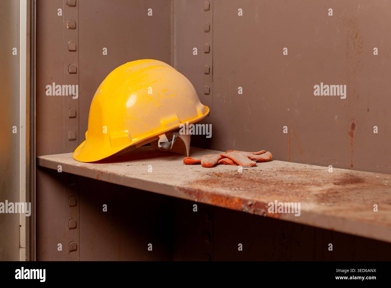 Elmetto giallo graffiato e guanti da lavoro arancioni usurati, dimenticati in un angolo di un armadietto metallico. Attrezzatura di sicurezza industriale vecchia, polverosa e trascurata. Foto Stock