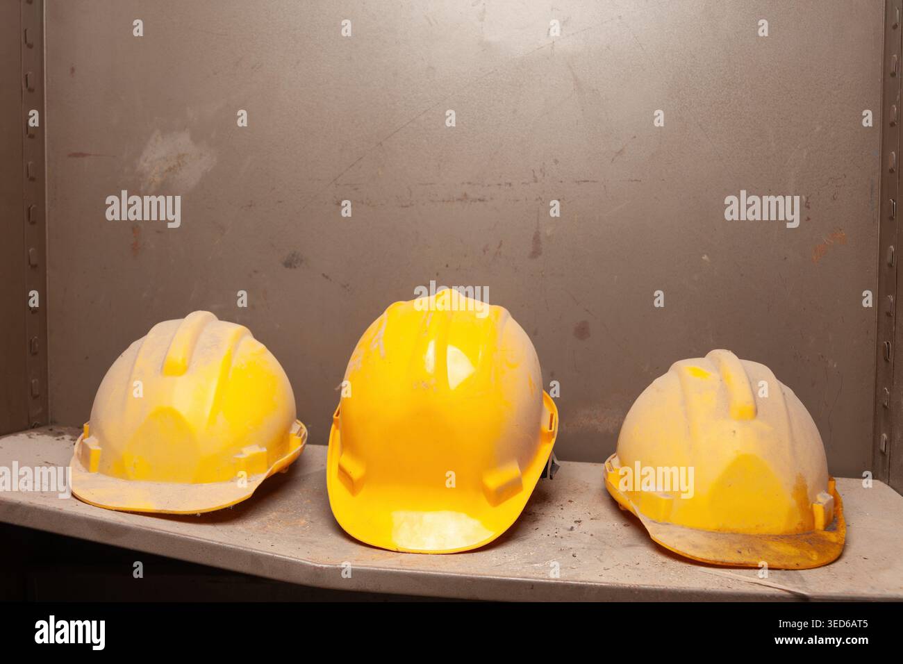 Tre cappelli rigidi gialli con diversi livelli di deterioramento sono posizionati in verticale in fila su un ripiano in metallo. Foto Stock