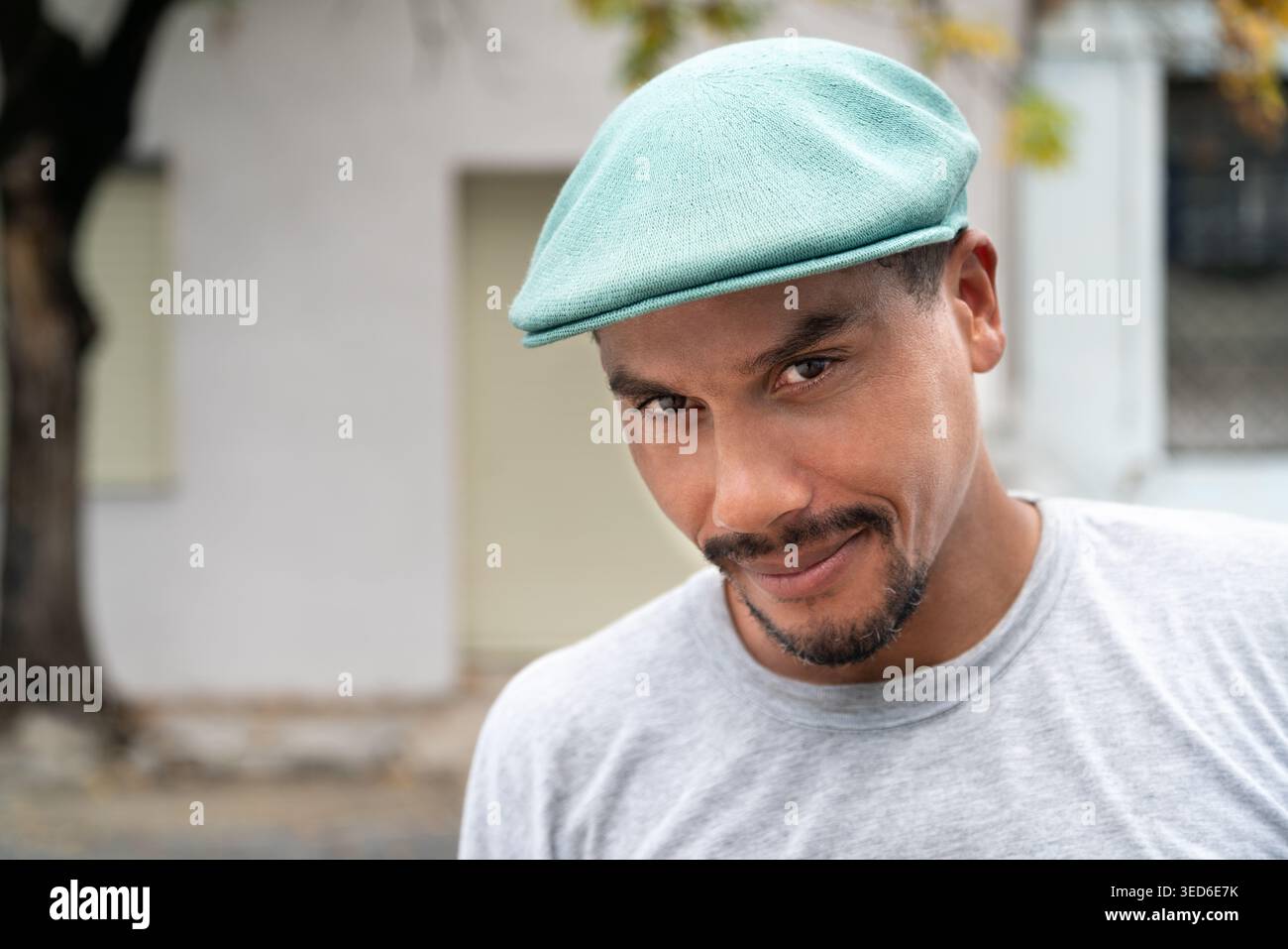 Un latino che indossa un cappello piatto che guarda la macchina fotografica, con un albero con foglie autunnali sullo sfondo. Copia spazio Foto Stock