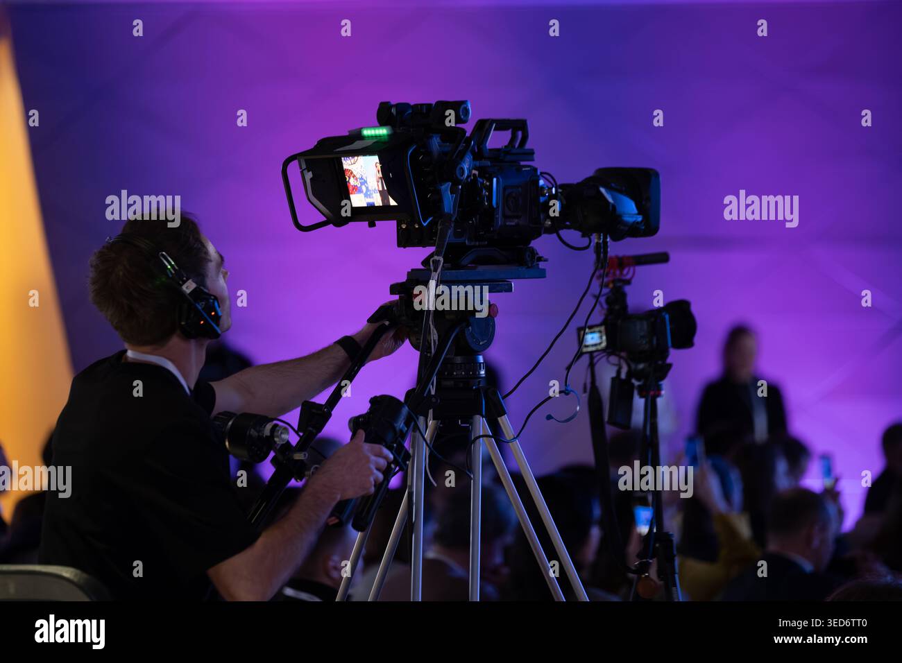 Cameraman che utilizza una videocamera professionale su treppiede durante un evento dal vivo, illuminazione viola del palco, pubblico in background. Foto Stock