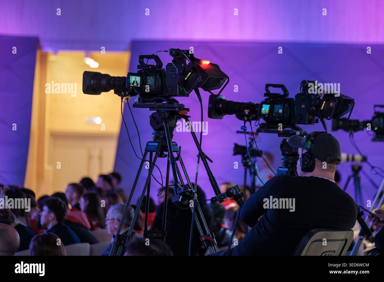 Più videocamere professionali e operatori di telecamere che riprendono conferenze dal vivo, illuminazione viola del palcoscenico, spettatori seduti, apparecchiature di produzione su treppiedi. Foto Stock