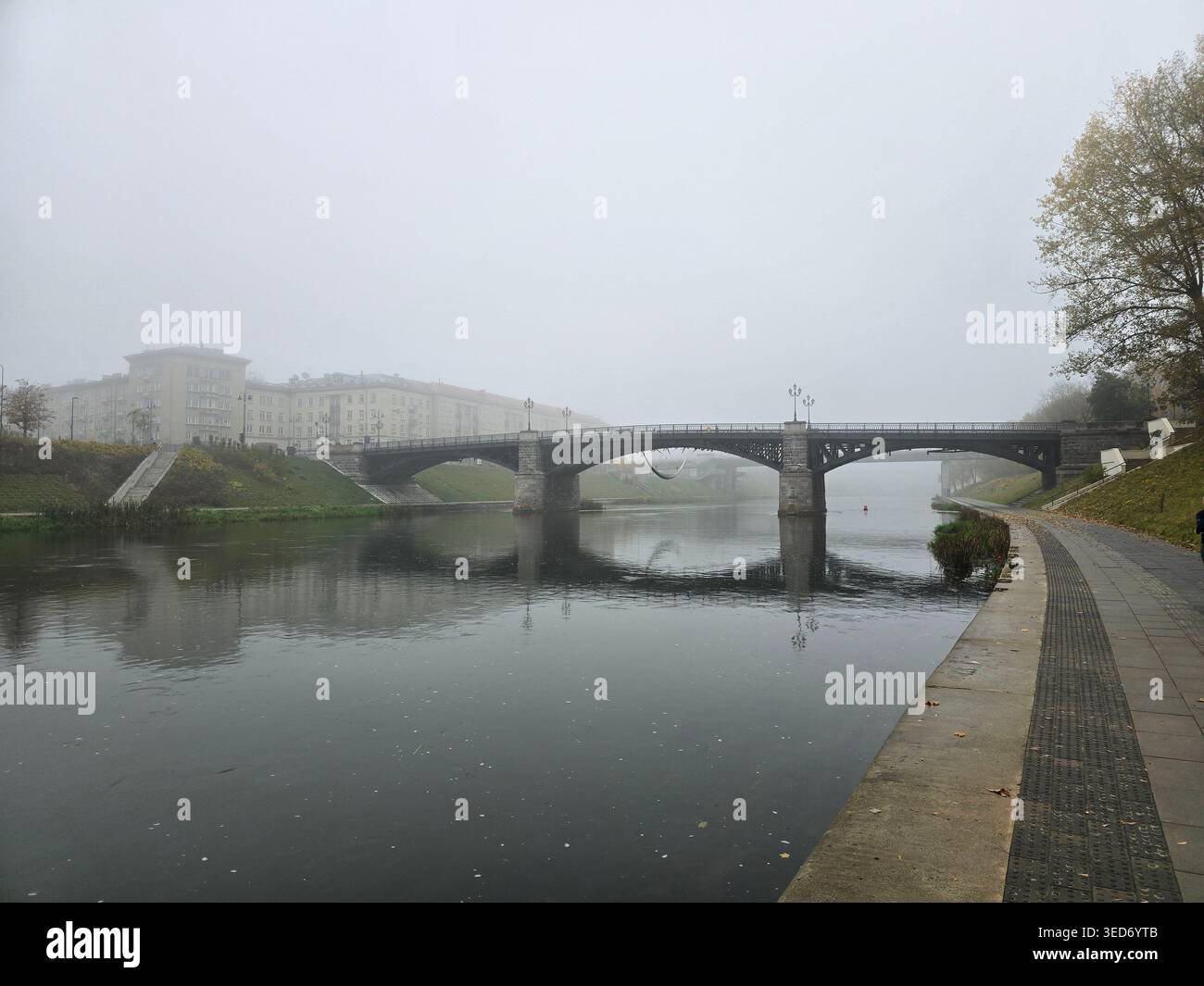 Un ponte di pietra attraversa un fiume calmo in una fitta nebbia mattutina lungo il fiume Neris a Vilnius, Lituania - Immagine stock catturata con smartphone