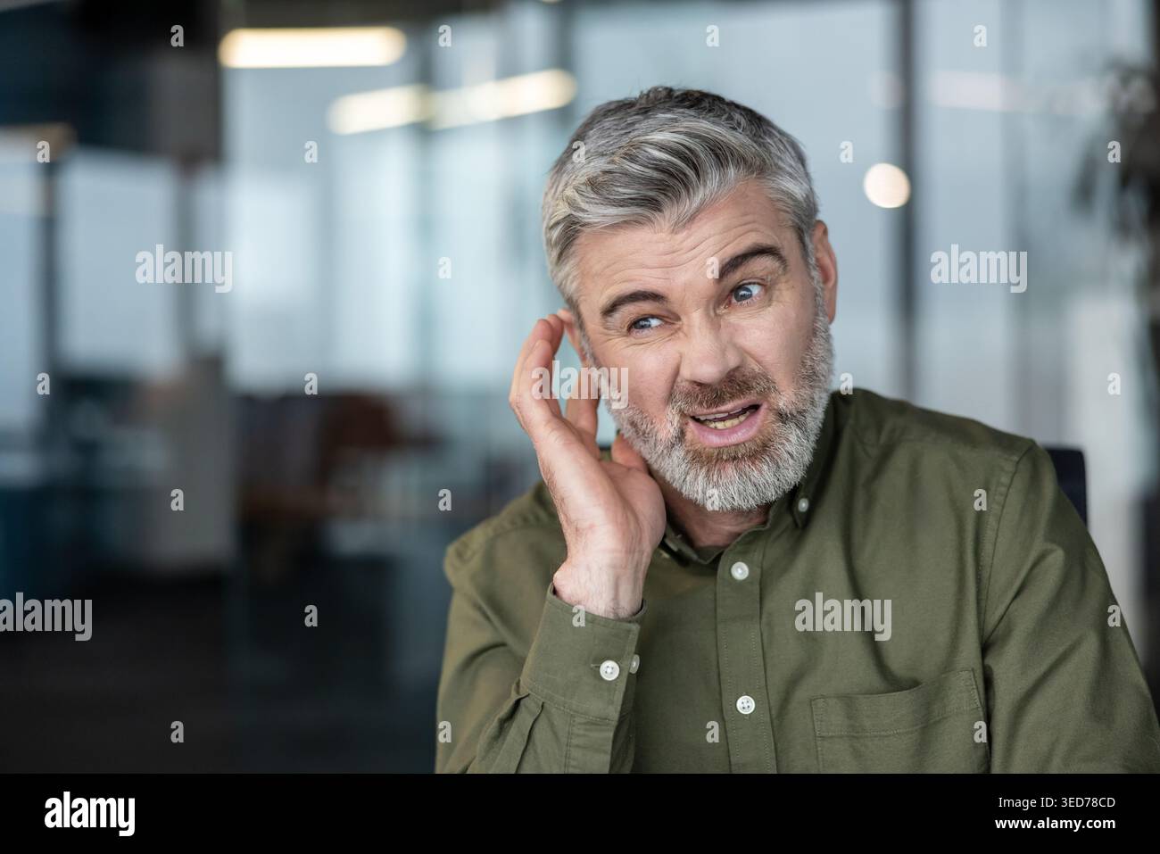 Uomo dai capelli grigi in un ufficio moderno che tocca l'orecchio e fatica a sentire, rappresentando concetti di perdita dell'udito, problemi di comunicazione e invecchiamento Foto Stock