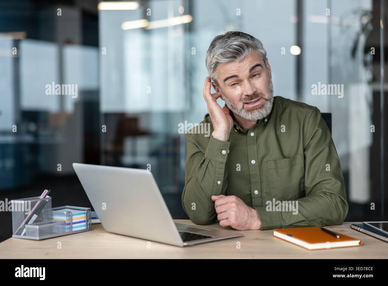 Uomo d'affari di mezza età che sembra confuso e si mette mano all'orecchio, ha problemi di udito o fatica a capire le informazioni mentre lavora con un notebook in un ufficio moderno Foto Stock