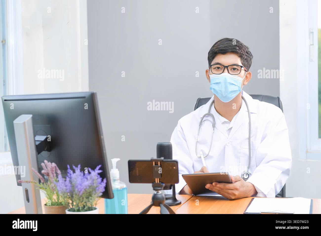 Ritratto di un medico seduto a una scrivania in un ufficio con dispositivi digitali assortiti che indossano una maschera per il viso e un cappotto da laboratorio Foto Stock
