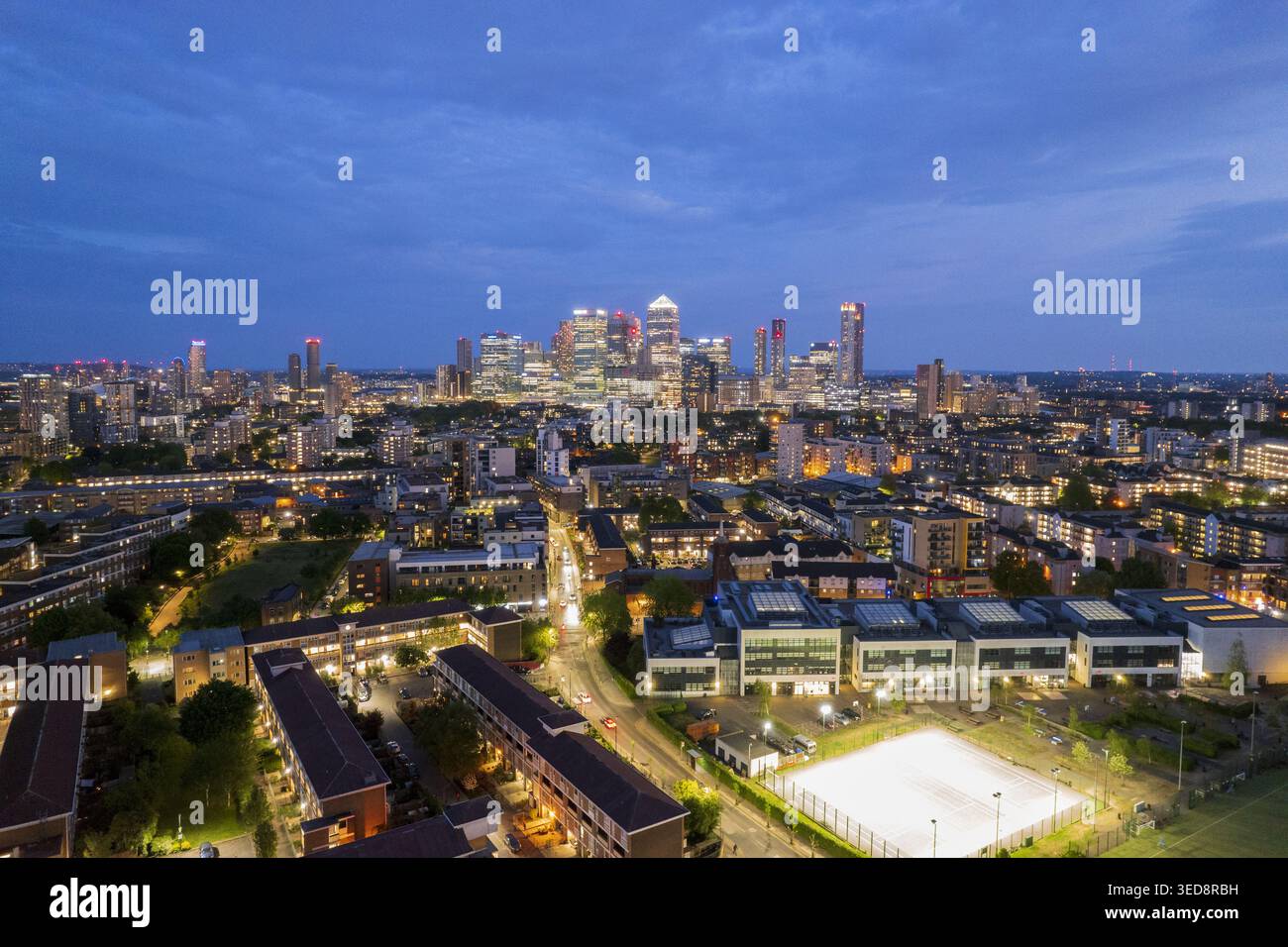 Vista aerea del quartiere di Mile End con blocchi residenziali e strade illuminate che conducono verso lo skyline del quartiere finanziario di Canary Wharf al crepuscolo L Foto Stock