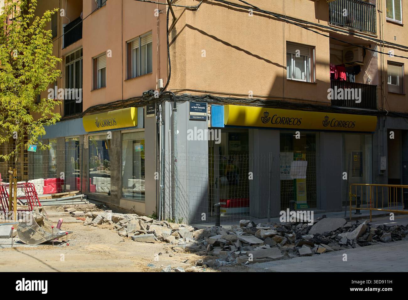Viladecans, Spagna - 4 maggio 2026: Un cartello giallo per uffici Correos su una facciata di un moderno edificio residenziale accanto a un cantiere con un carico pesante Foto Stock