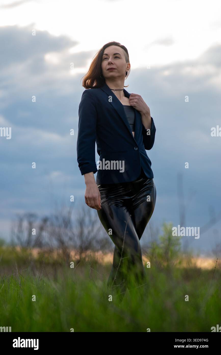 Ritratto di una donna sicura di sé che indossa un blazer blu navy e leggings neri lucenti, in posa in un campo erboso sotto un cielo nuvoloso. Alla moda, svuotare Foto Stock