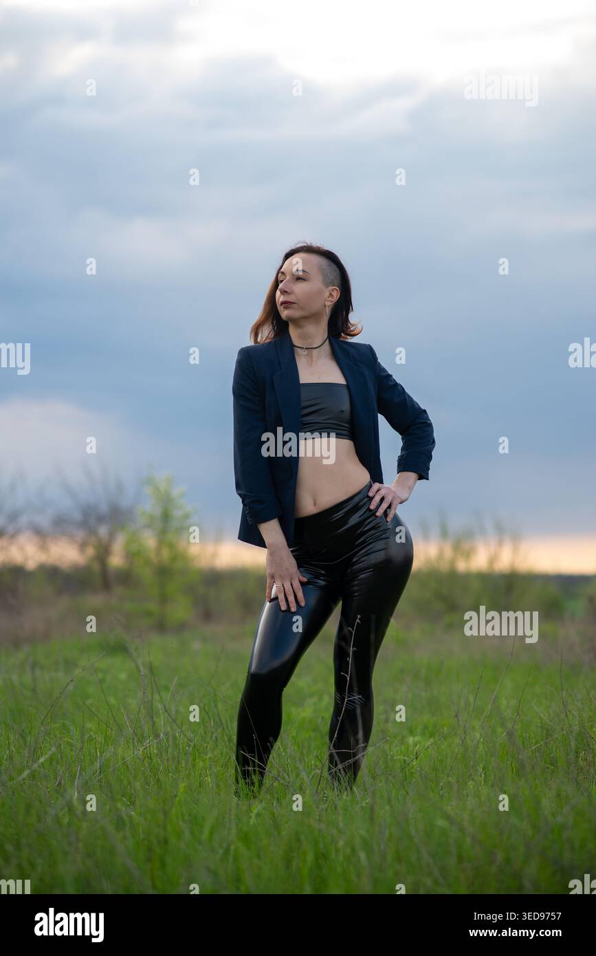 Una donna elegante con sottosquadro posa in sicurezza in un campo erboso al crepuscolo, indossando un blazer, un top raccolto e leggings luccicanti. Moody Sky e paesaggio naturale Foto Stock