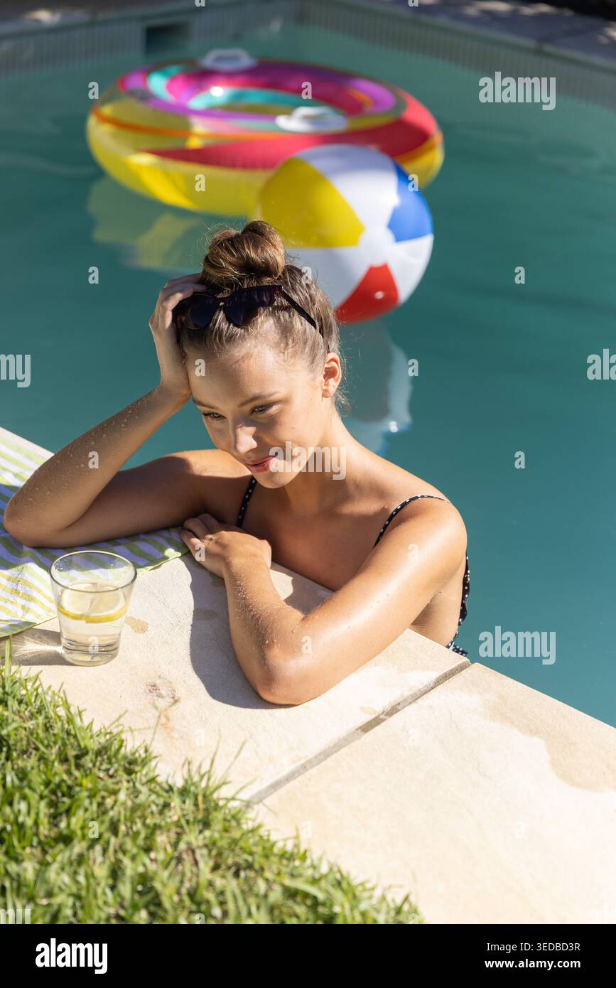 Donna che si appoggia al costume da bagno nella piscina del cortile, bicchiere accanto all'asciugamano, anello e palla galleggiante Foto Stock