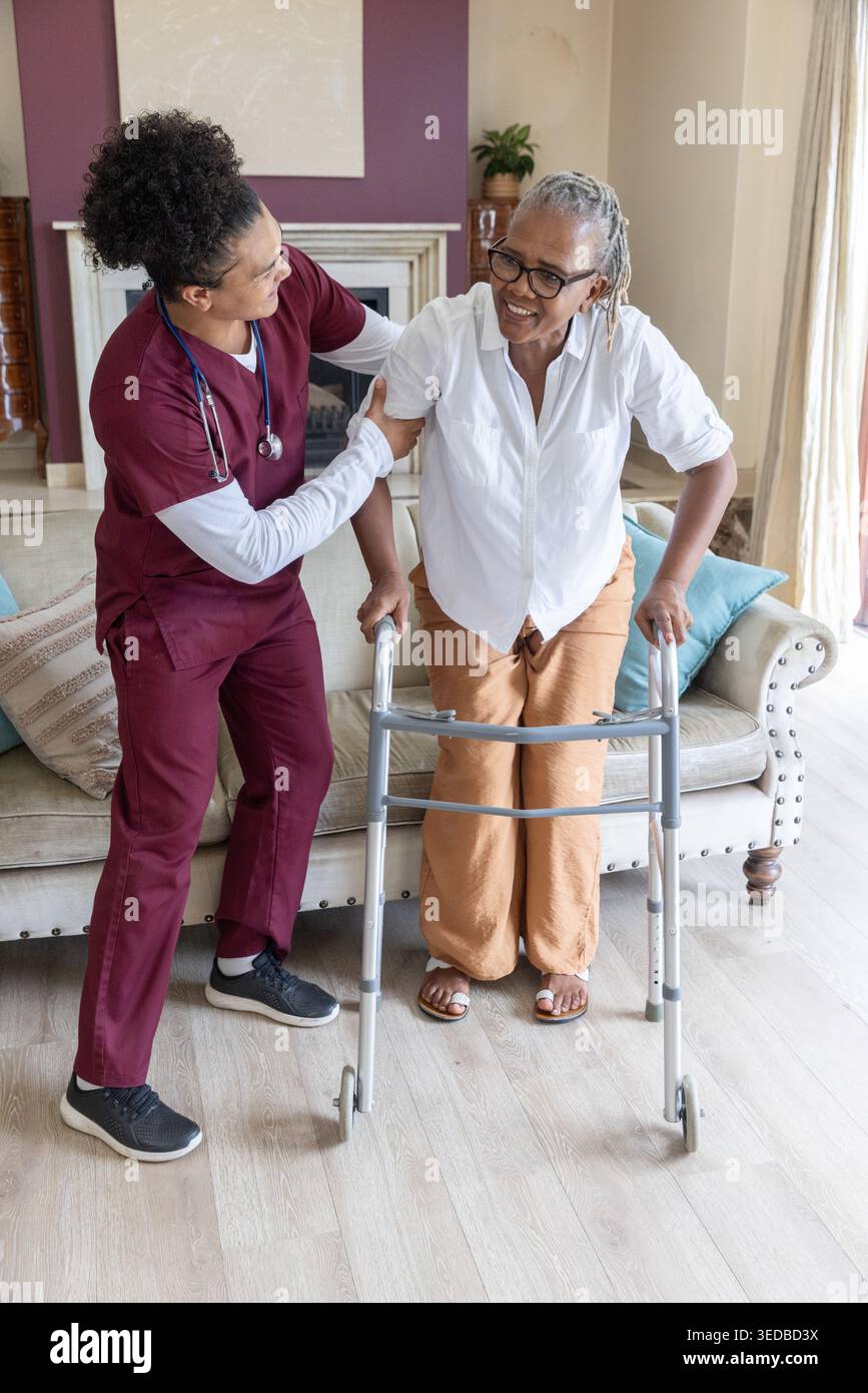 Caregiver in scrub di colore marrone che sostiene una donna afroamericana anziana che usa un passeggio in salotto Foto Stock