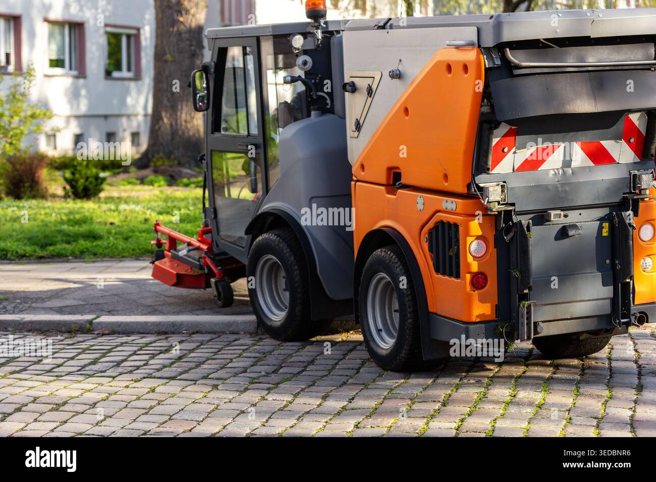 L'attrezzatura di falciatura della macchina per la manutenzione municipale opera su marciapiedi pavimentazioni in ciottoli ambiente di parchi urbani Germania. Il veicolo compatto mantiene l'erba Foto Stock