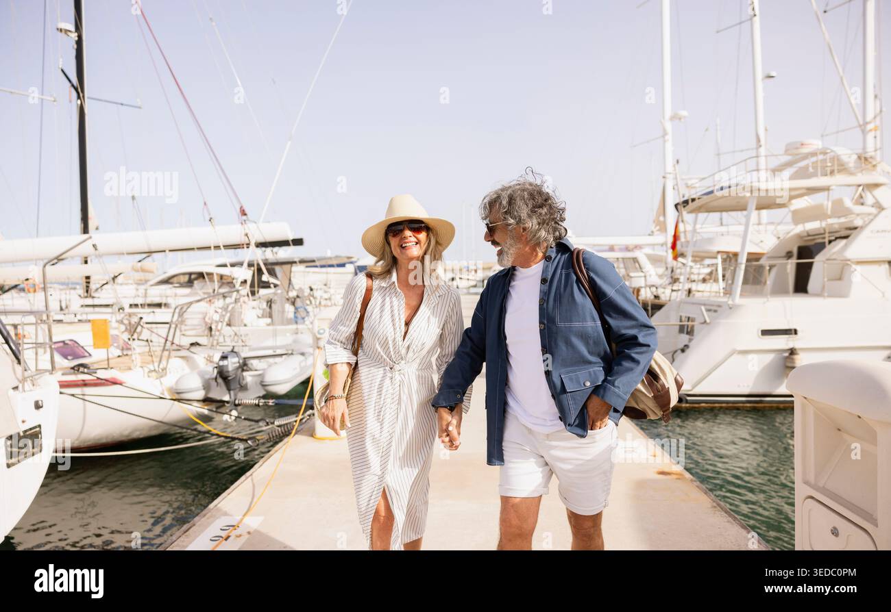 Felice coppia di anziani che si tiene per mano, godendosi una vacanza estiva passeggiando lungo il porto Foto Stock