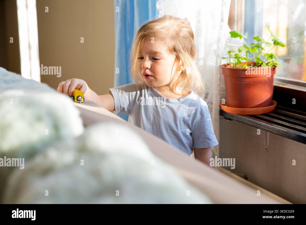Bambino concentrato che gioca con un'auto di legno gialla vicino a una finestra, gioco sensoriale, umanità disordinata, luce mattutina, esperienza tattile, belle capacità motorie Foto Stock