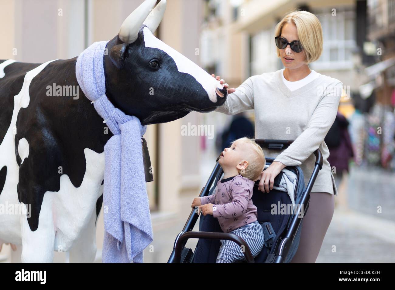 CuriouMother e Little Son esplorano le strade della città e scoprono le divertenti sculture di una mucca per bambini in un passeggino che guarda una statua di mucca di fronte a un negozio in Spagna. Foto Stock