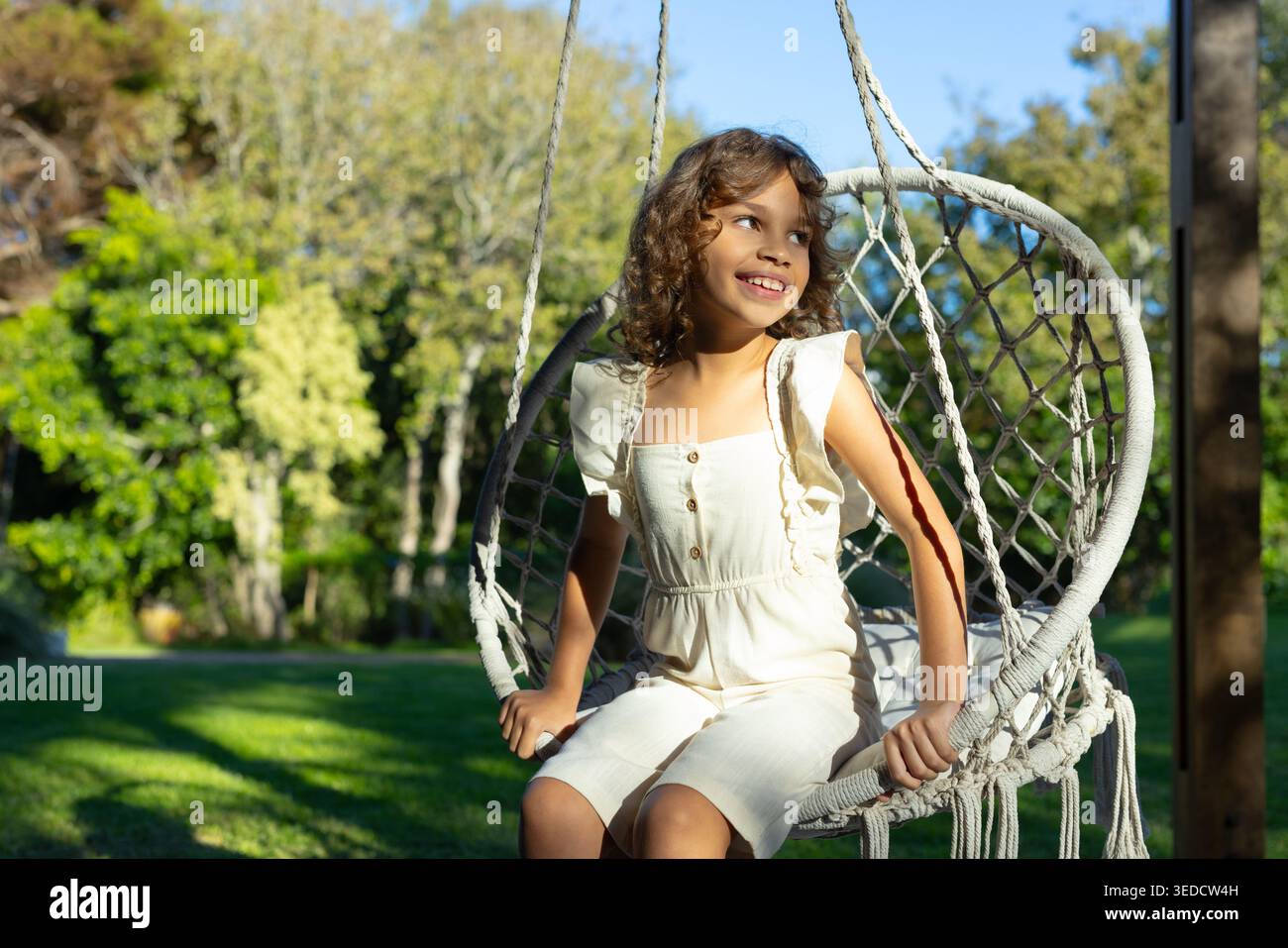 Preadolescente afroamericana seduta su una sedia in macrame in giardino tenendo la corda in una crema romper Foto Stock