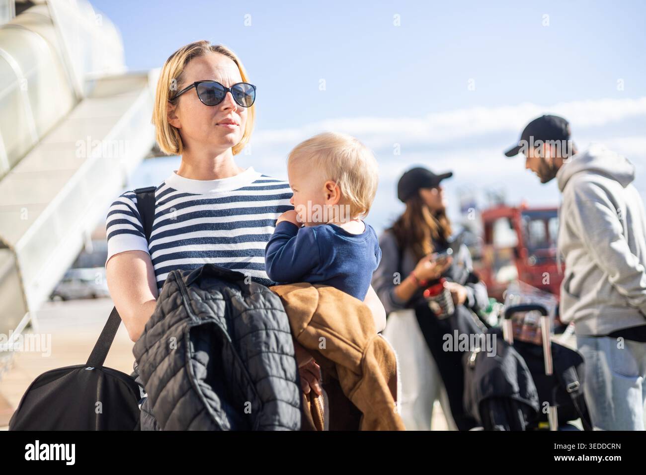 La madre viaggia con il suo bambino. La mamma tiene in mano la borsa da viaggio e il suo bambino mentre fa la fila per l'autobus di fronte alla stazione del terminal dell'aeroporto Foto Stock