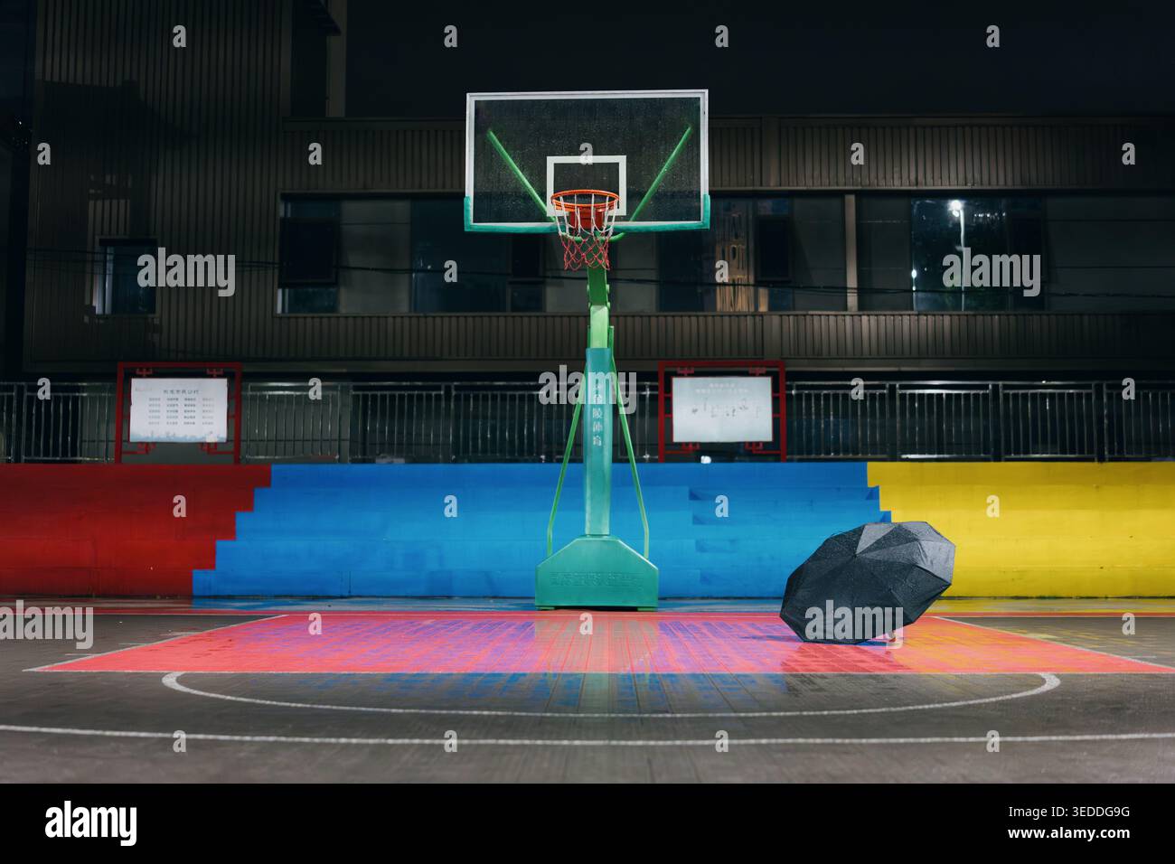 Campo da pallacanestro vuoto di notte con ombrello solitario appoggiato su un pavimento colorato e sport urbani Foto Stock
