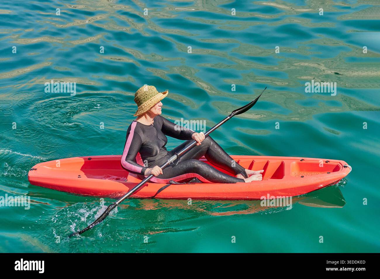 Donna matura che si diverte a pagaiare in kayak rosso brillante. Indossando muta e cappello di paglia, esplora le calme acque color smeraldo. Stile di vita attivo e guarigione Foto Stock