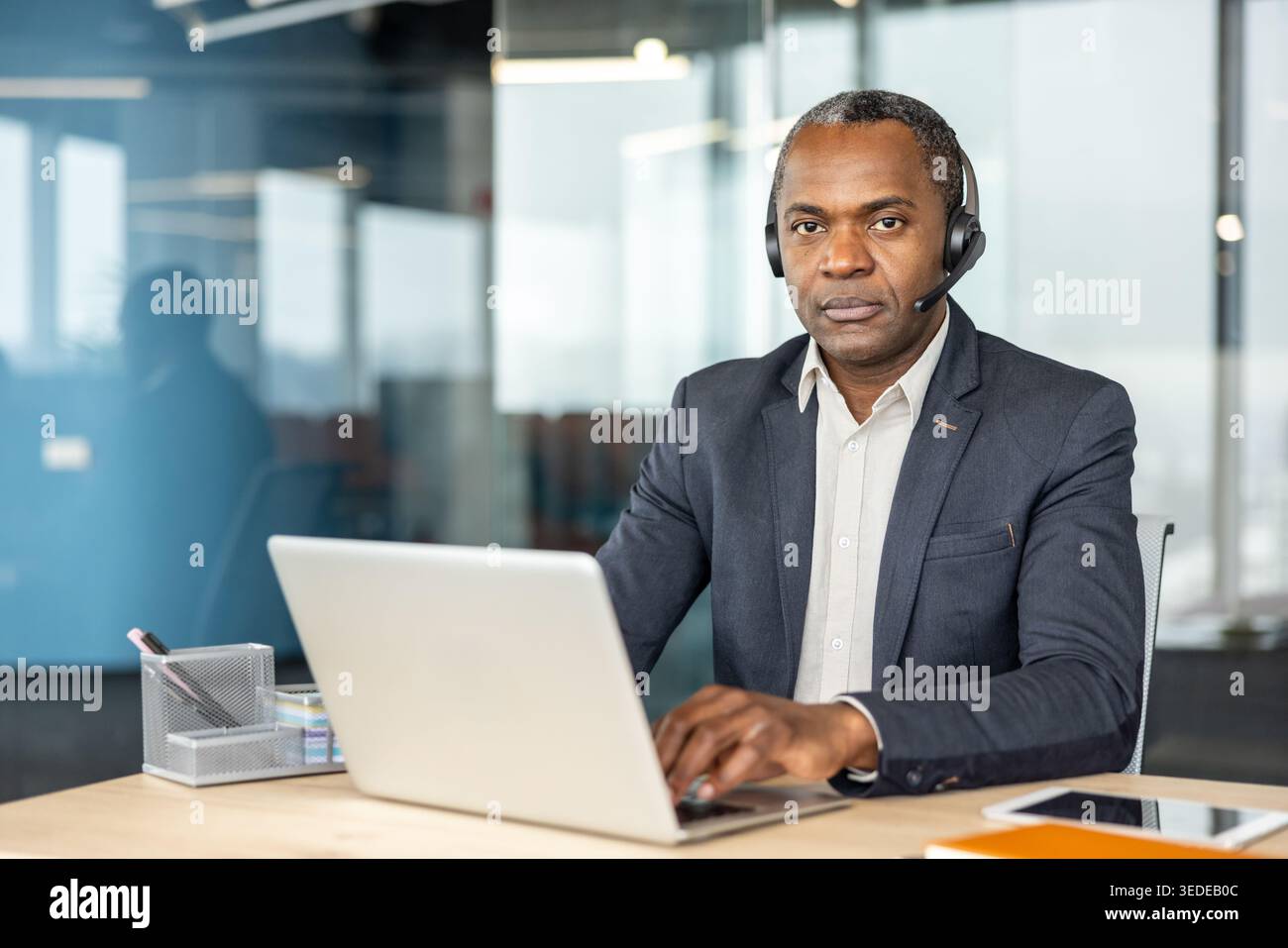 Un rappresentante del servizio clienti professionale di sesso maschile che indossa una cuffia e digita un laptop, impegnandosi attivamente nella comunicazione e nell'assistenza in un ambiente aziendale moderno Foto Stock